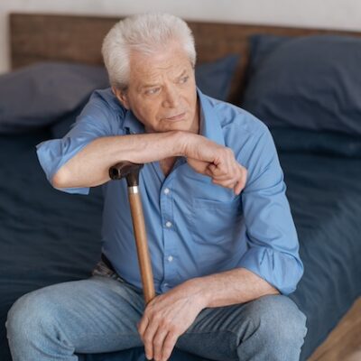 An elderly man sits on his bed with his walking stick.