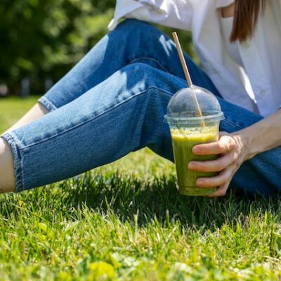 A young girl is sitting in the park on the grass and drinking a green smoothie.